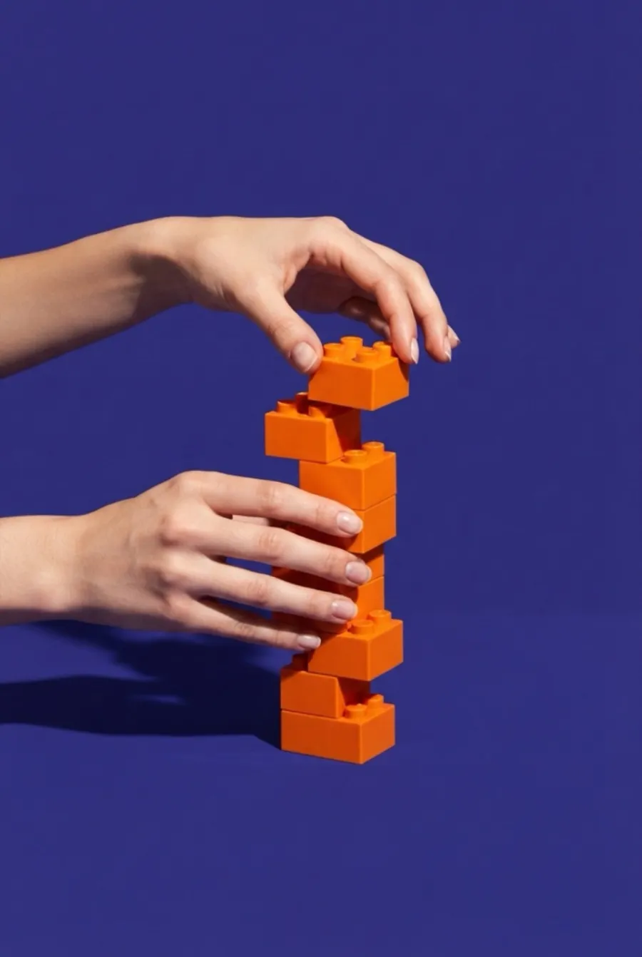 Zwei Hände stapeln leuchtend orange Bausteine vor lila Hintergrund